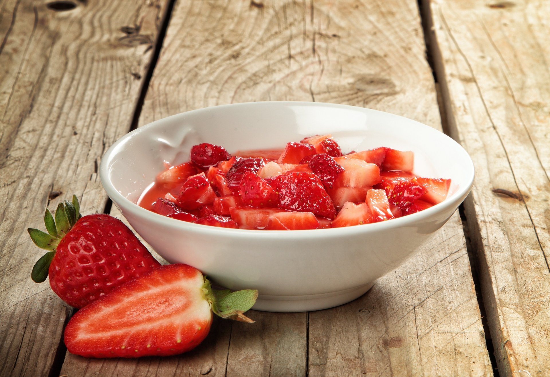 A white bowl filled with chopped strawberries on a rustic wooden surface, accompanied by whole and sliced strawberries, featured as an HD PC desktop wallpaper background.