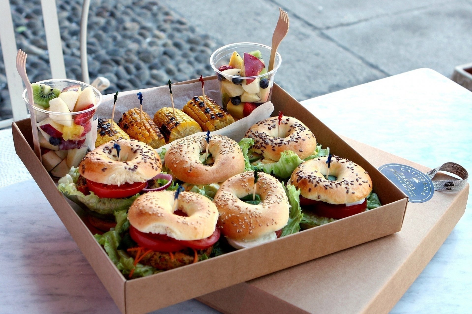 HD desktop wallpaper showing a box of fresh sandwiches with lettuce and tomato, accompanied by grilled corn and fruit cups on a white table.