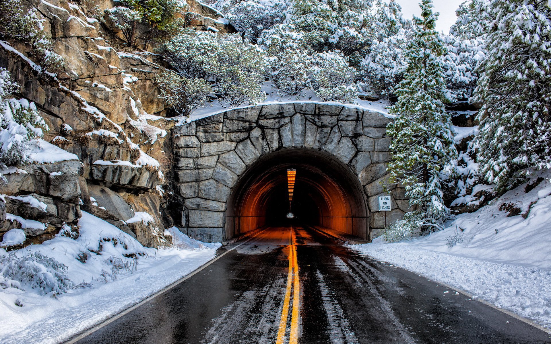HD PC desktop wallpaper and background of a man-made stone tunnel carved into snowy rock, wet road with yellow centerlines leading to a warm, glowing interior.