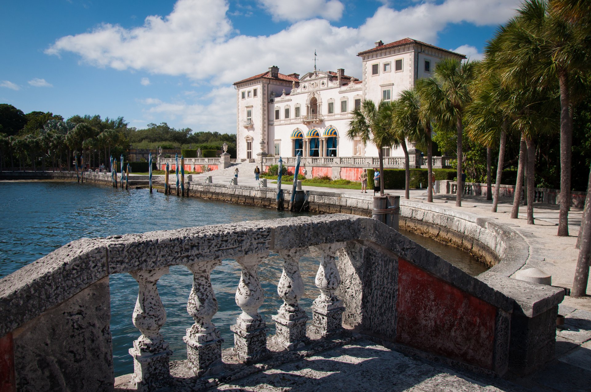 A stunning view of Villa Vizcaya in Florida, showcasing its elegant architecture alongside the serene water, framed by palm trees and a decorative stone railing.
