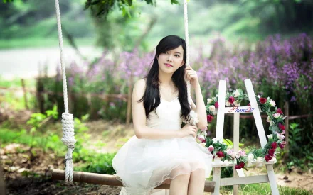 An Asian woman in a white dress sits on a swing in a serene garden, surrounded by lush greenery and purple flowers. The HD desktop wallpaper captures a peaceful, natural background.