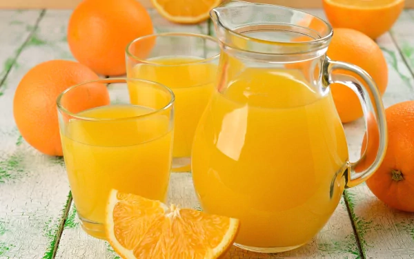 4K Ultra HD desktop wallpaper featuring a clear glass pitcher and two glasses filled with fresh orange juice surrounded by whole and sliced bright orange oranges.