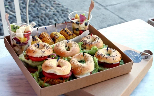 HD desktop wallpaper showing a box of fresh sandwiches with lettuce and tomato, accompanied by grilled corn and fruit cups on a white table.