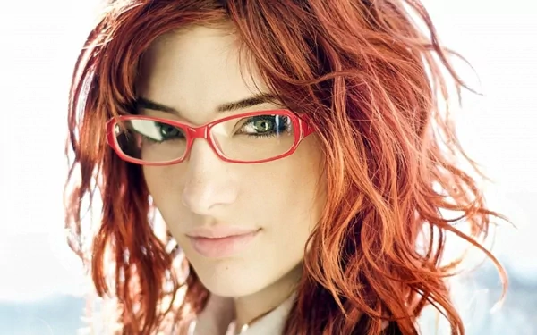HD desktop wallpaper featuring celebrity Susan Coffey with red hair and red glasses against a bright, softly blurred background.