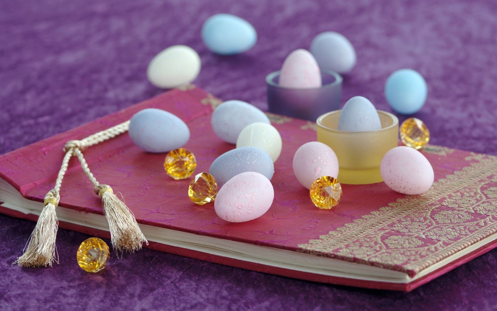 A vibrant Easter-themed desktop wallpaper featuring pastel-colored eggs and decorative gems arranged on a purple fabric background and a decorative book.