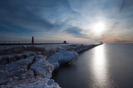 HD photography of a serene coastline at sunset, featuring icy rocks and a distant lighthouse under a cloudy sky, captured as a desktop wallpaper and background.