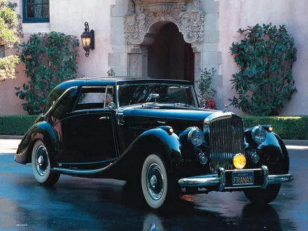 A classic black Bentley parked in front of an elegant building, surrounded by lush greenery, showcasing its timeless design in this HD desktop wallpaper.