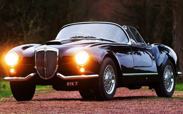 HD desktop wallpaper featuring a classic Lancia Aurelia B24 Spider with headlights on, parked on a rustic ground against a dark forest backdrop.