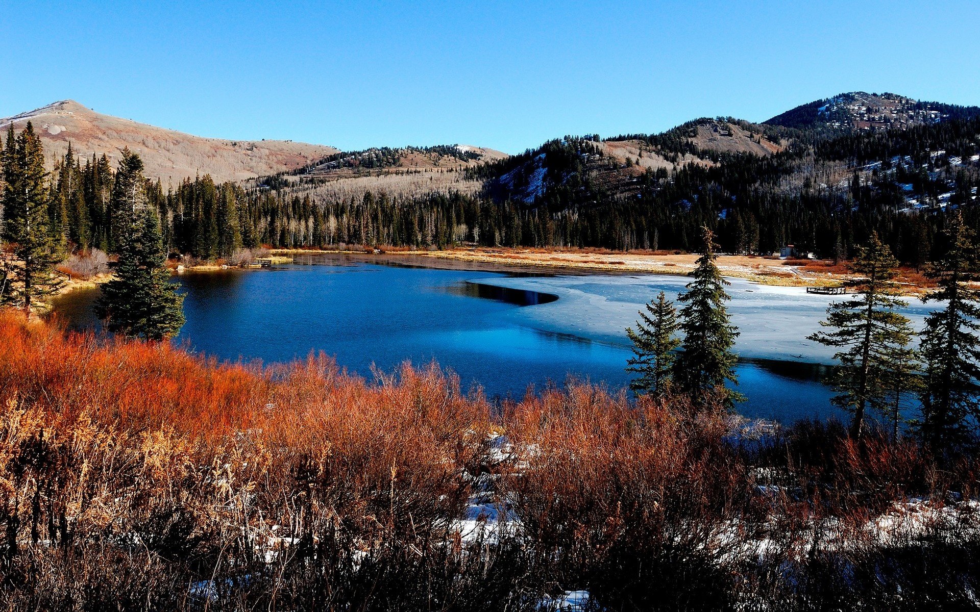 HD desktop wallpaper showcasing a serene lake surrounded by autumn foliage and evergreen trees, set against a backdrop of mountainous landscape and clear blue sky.