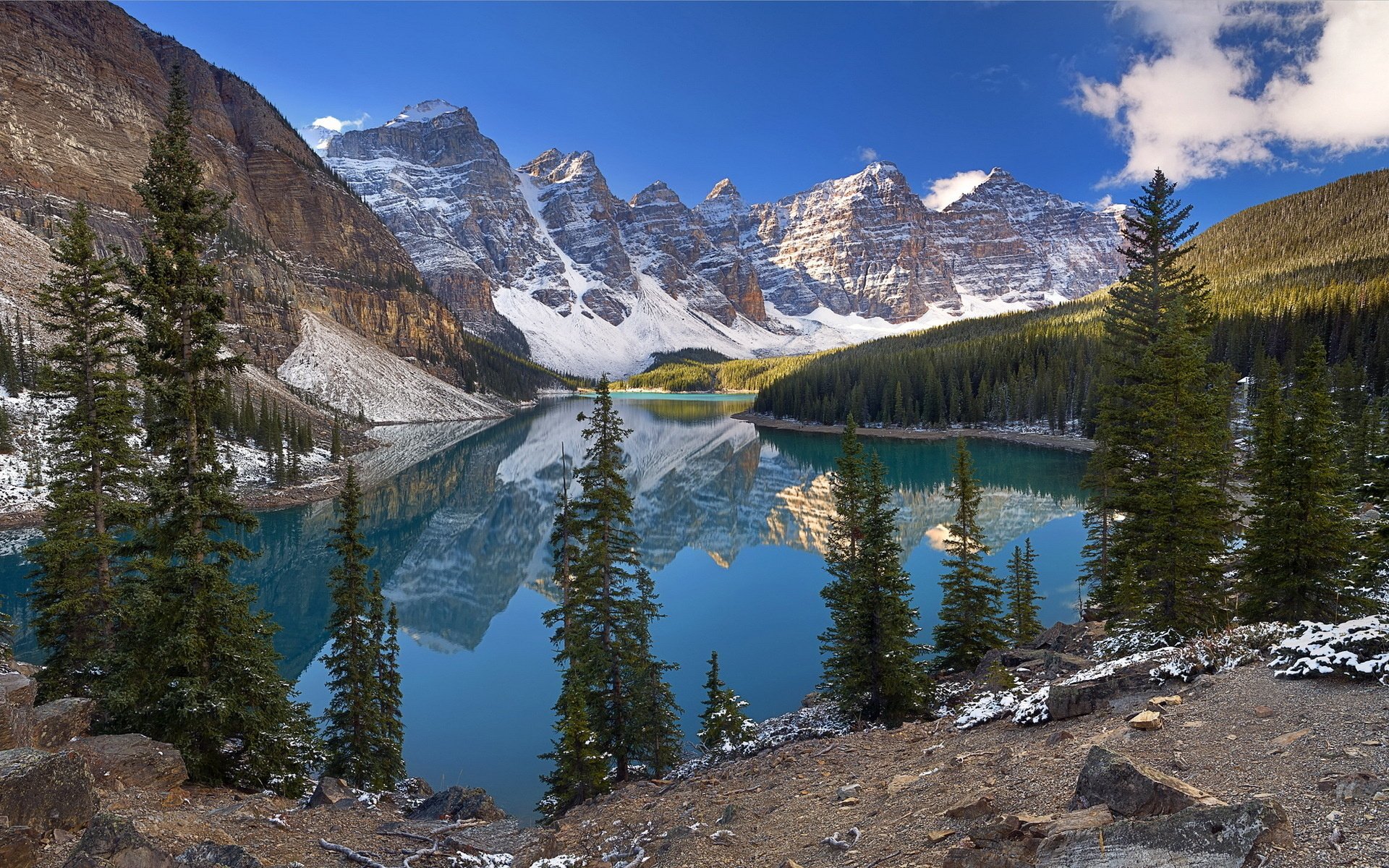HD PC desktop wallpaper: a nature scene of an alpine turquoise lake reflecting snow-capped peaks and evergreens beneath a blue sky, rocky foreground framing the mirror-like reflection.