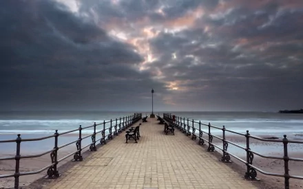 HD desktop wallpaper of a man-made pier extending into calm waters under a dramatic, cloudy sky with benches lining the walkway.
