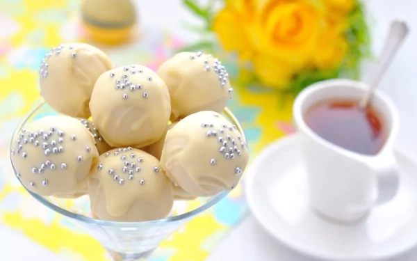 A delightful display of white chocolate truffles with silver sprinkles in a glass bowl, accompanied by a cup of tea and yellow roses, creating an inviting food-themed wallpaper.