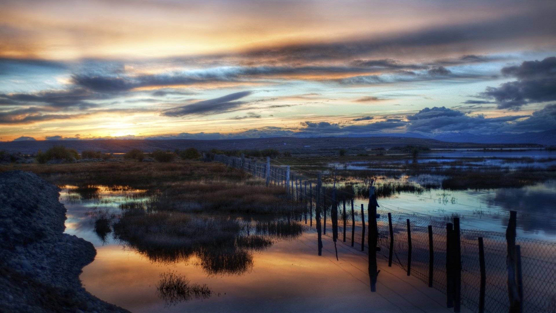 A stunning reflection of a colorful sunset over calm waters, framed by rustic wooden posts and lush grasslands, creating a serene HD desktop wallpaper and background.