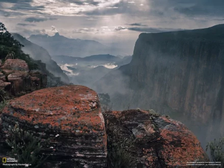 HD PC desktop wallpaper: mist-shrouded canyon with red-rock ledge in foreground, sheer cliffs and distant mesas under a dramatic cloudy sky.