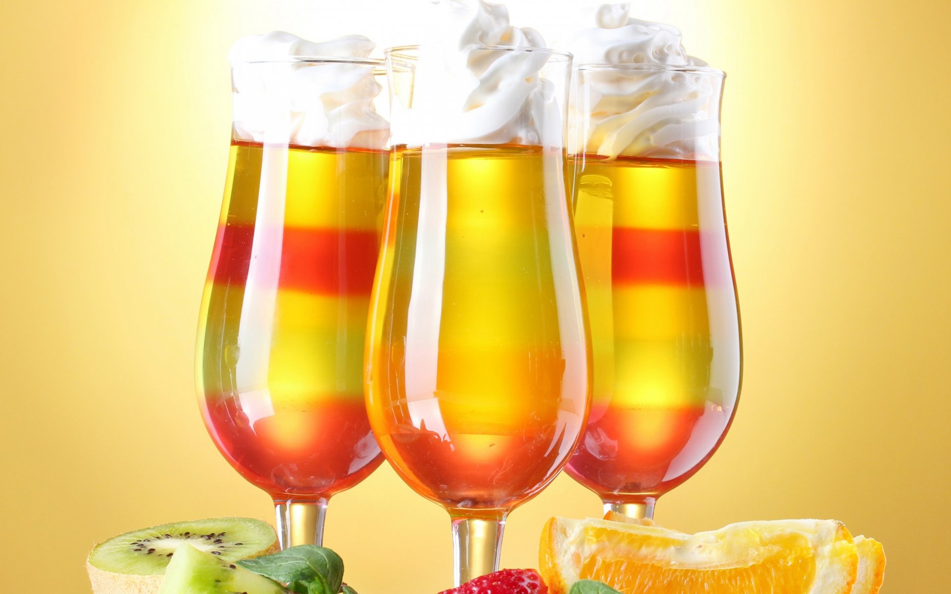 HD PC desktop wallpaper featuring three vibrant layered cocktails garnished with whipped cream, surrounded by fresh fruit slices including kiwi, strawberry, and orange.