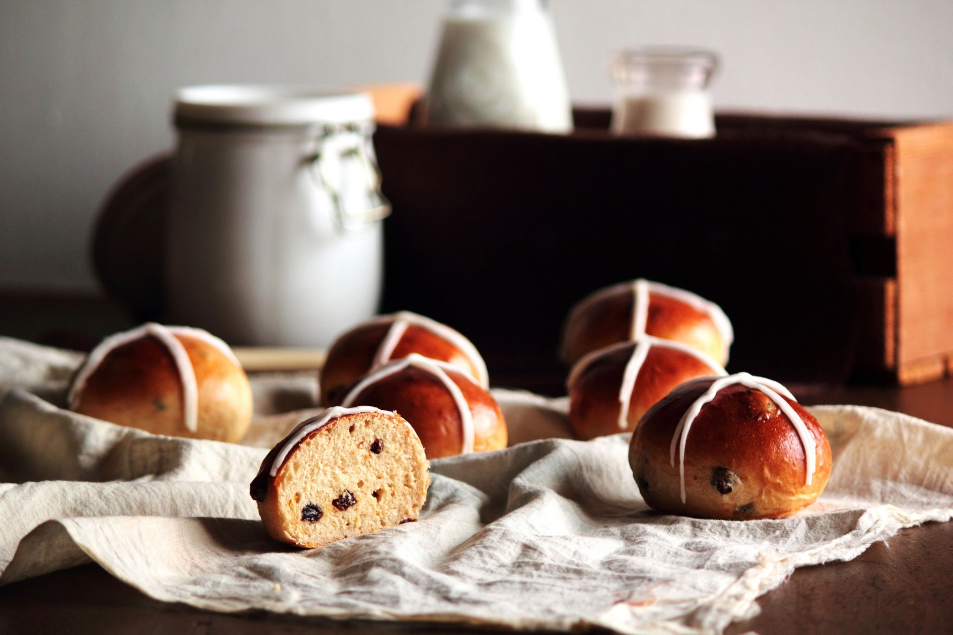 HD PC desktop wallpaper showing freshly baked hot cross buns with one cut open, set on a cloth with jars blurred in the background, highlighting food and baking.