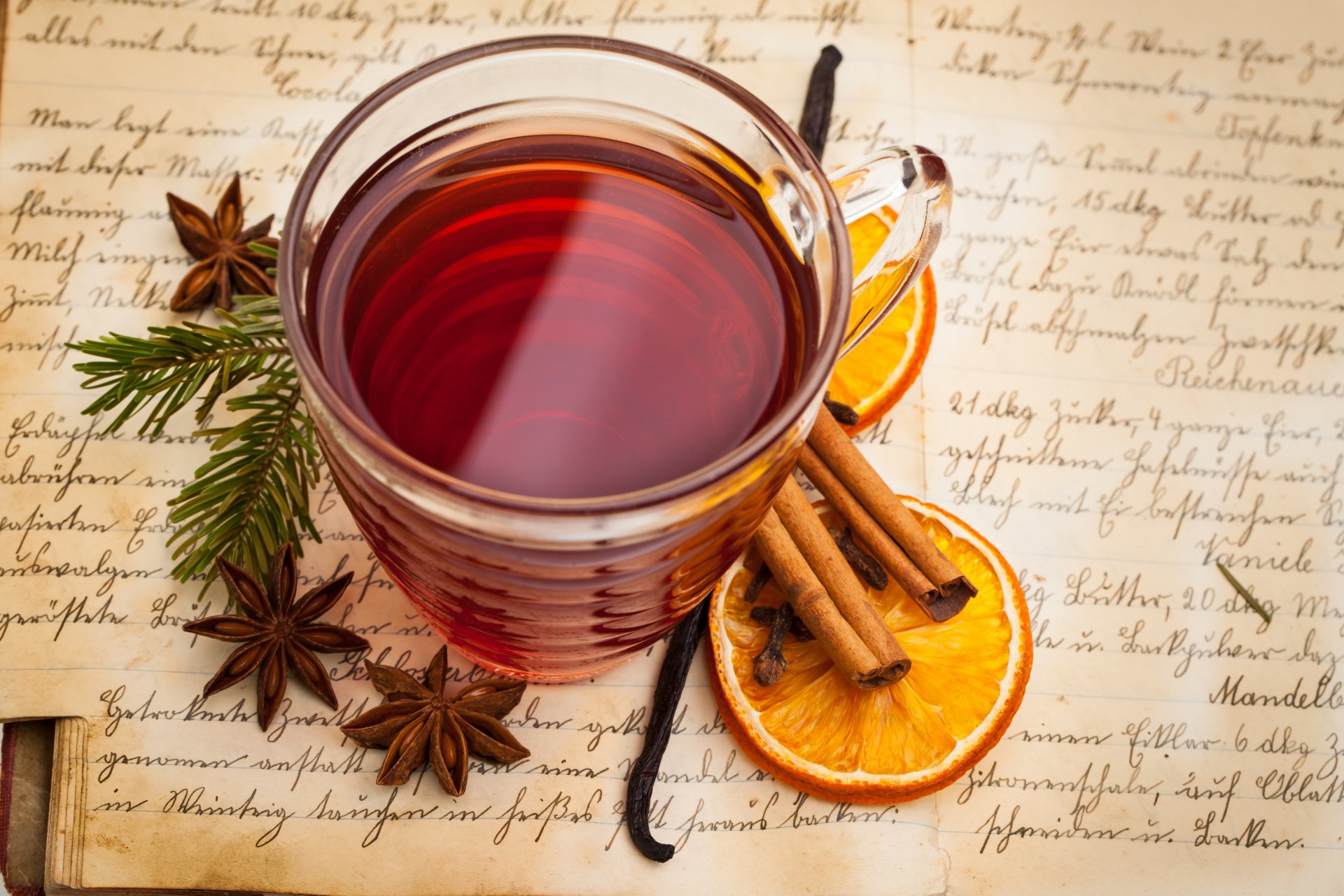 A 4K Ultra HD desktop wallpaper featuring a clear glass cup of tea surrounded by cinnamon sticks, dried orange slices, star anise, and pine sprigs on aged handwritten paper.