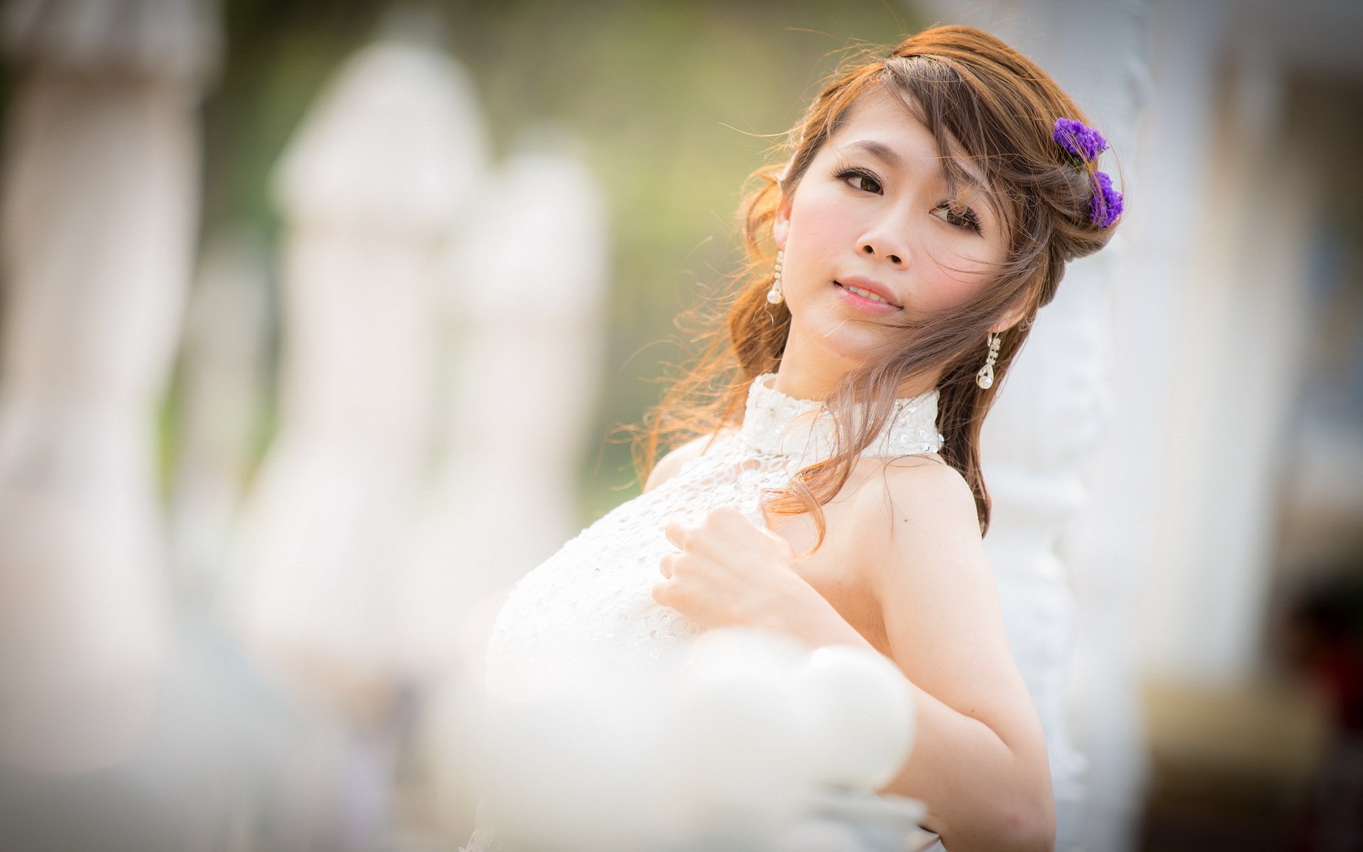 HD wallpaper with an Asian woman in a serene outdoor setting, her hair gently tousled and adorned with a purple flower, wearing a white lace dress.