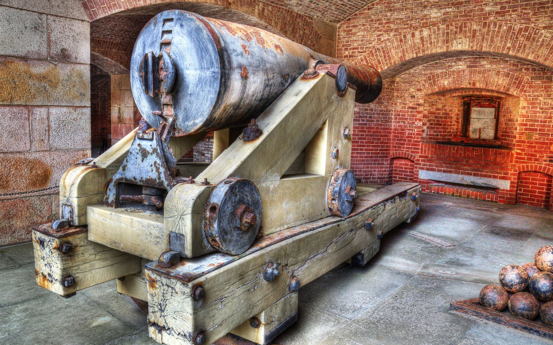 HD desktop wallpaper showing a man-made historic cannon inside a brick-walled fortress room with cannonballs stacked nearby.