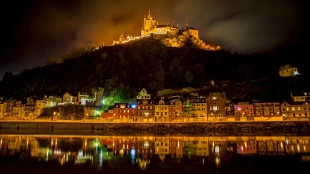 Night view of Cochem, Germany, with illuminated castle on a hill, glowing town lights, and their reflections on the calm lake below.