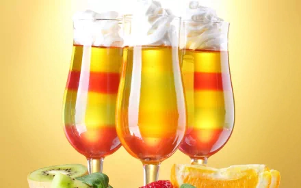 HD PC desktop wallpaper featuring three vibrant layered cocktails garnished with whipped cream, surrounded by fresh fruit slices including kiwi, strawberry, and orange.