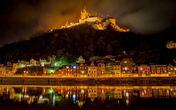 Night view of Cochem, Germany, with illuminated castle on a hill, glowing town lights, and their reflections on the calm lake below.