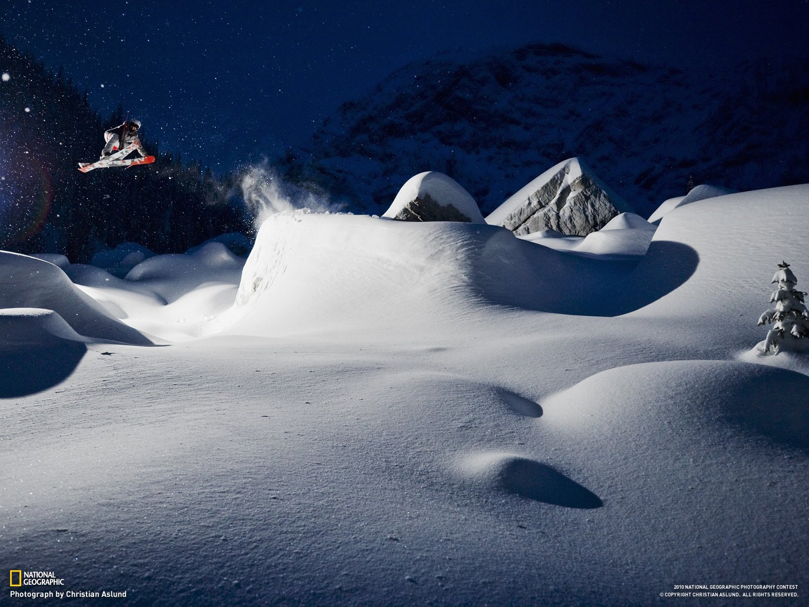 HD PC desktop wallpaper showing a skier mid-air performing a jump over snow-covered terrain under a dark, snowy night sky, capturing the thrill of skiing and sports.