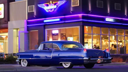 A classic 1956 Cadillac parked outside a retro diner at night, captured in a vibrant HD PC desktop wallpaper showcasing neon lights and vintage charm.