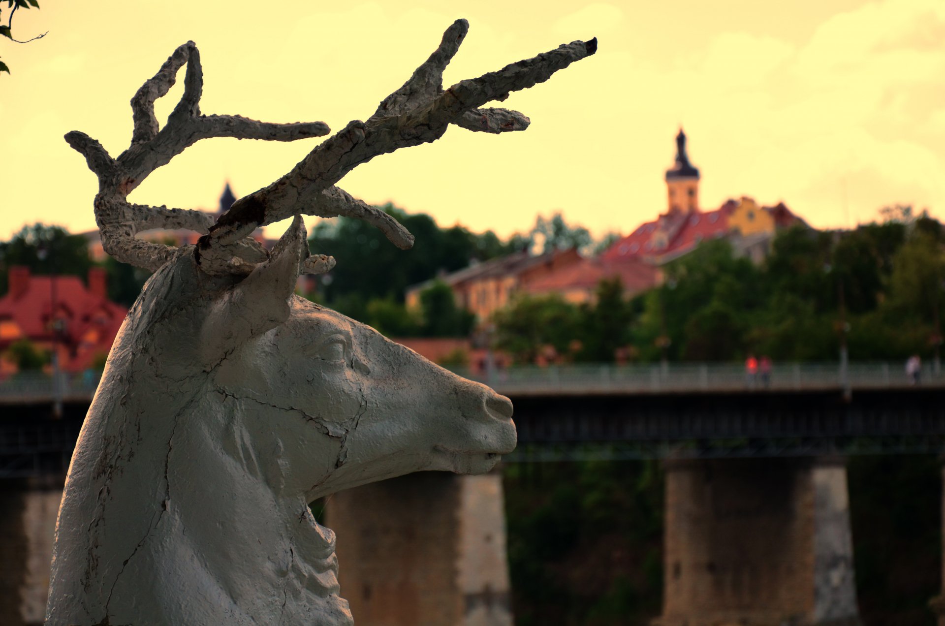 Man-made statue of a stag with branching antlers in the foreground, river bridge and town steeple at sunset — 4K Ultra HD PC desktop wallpaper and background.