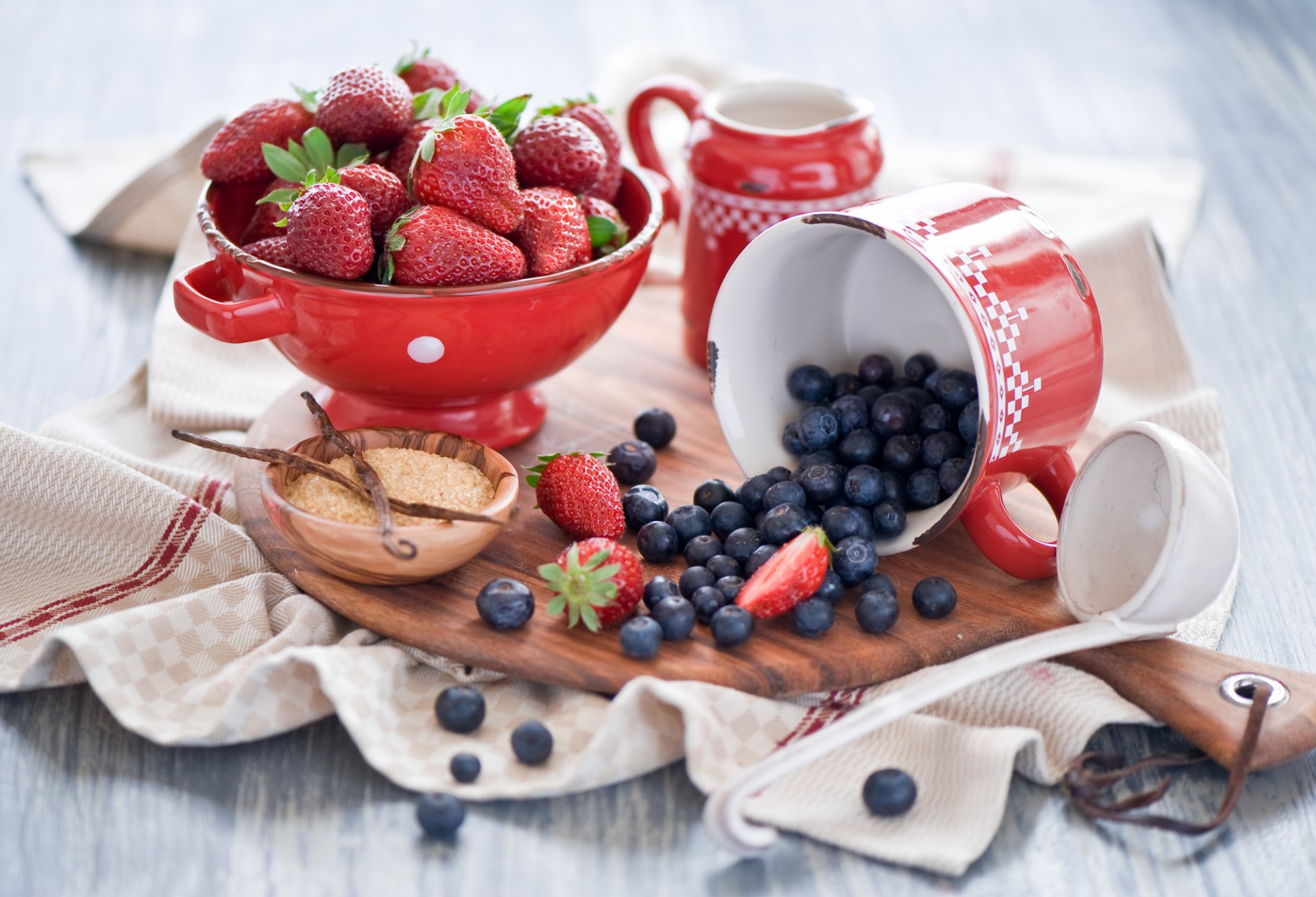 HD desktop wallpaper featuring a still life of fresh strawberries in a red bowl and blueberries spilling from a tipped red mug on a wooden board with rustic table linens.