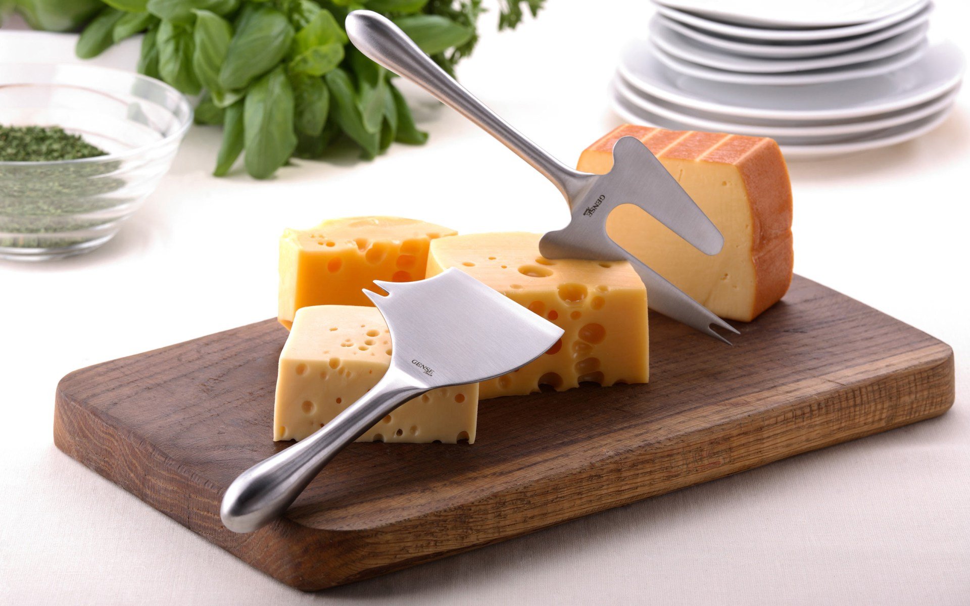 HD desktop wallpaper featuring assorted cheese wedges on a wooden board with cheese slicers, plates stacked in the background, and fresh herbs.