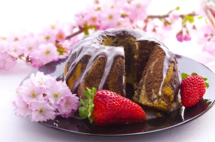 A delicious cake drizzled with icing sits on a black plate, accompanied by fresh strawberries and surrounded by delicate cherry blossoms—an inviting scene for a baking-themed background.