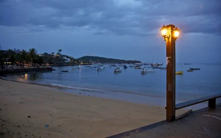 A tranquil beach scene at dusk, featuring a glowing lamp post beside the shore, with boats dotting the calm water in the background, creating a serene atmosphere.