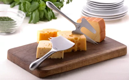 HD desktop wallpaper featuring assorted cheese wedges on a wooden board with cheese slicers, plates stacked in the background, and fresh herbs.