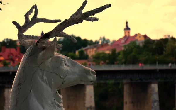 Man-made statue of a stag with branching antlers in the foreground, river bridge and town steeple at sunset — 4K Ultra HD PC desktop wallpaper and background.