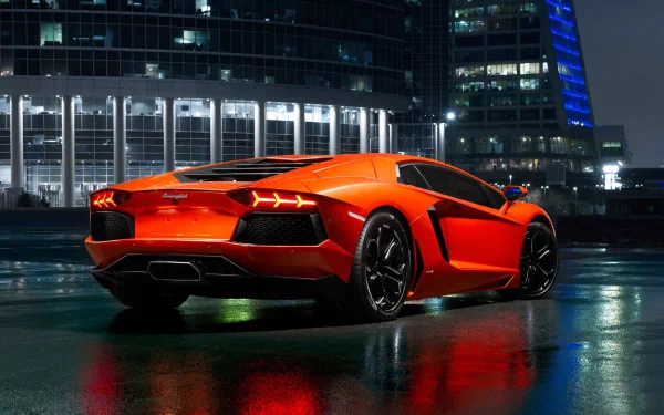 HD desktop wallpaper featuring an orange Lamborghini Aventador parked at night against a backdrop of illuminated modern buildings. The sleek, high-performance sports car glistens under the city lights.
