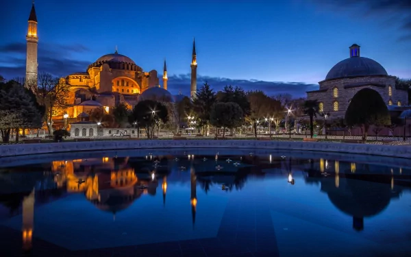 HD desktop wallpaper of Hagia Sophia at night, beautifully illuminated with its reflection in a calm, mirror-like pool in the foreground. The historical and religious architecture stands majestically against the twilight sky.