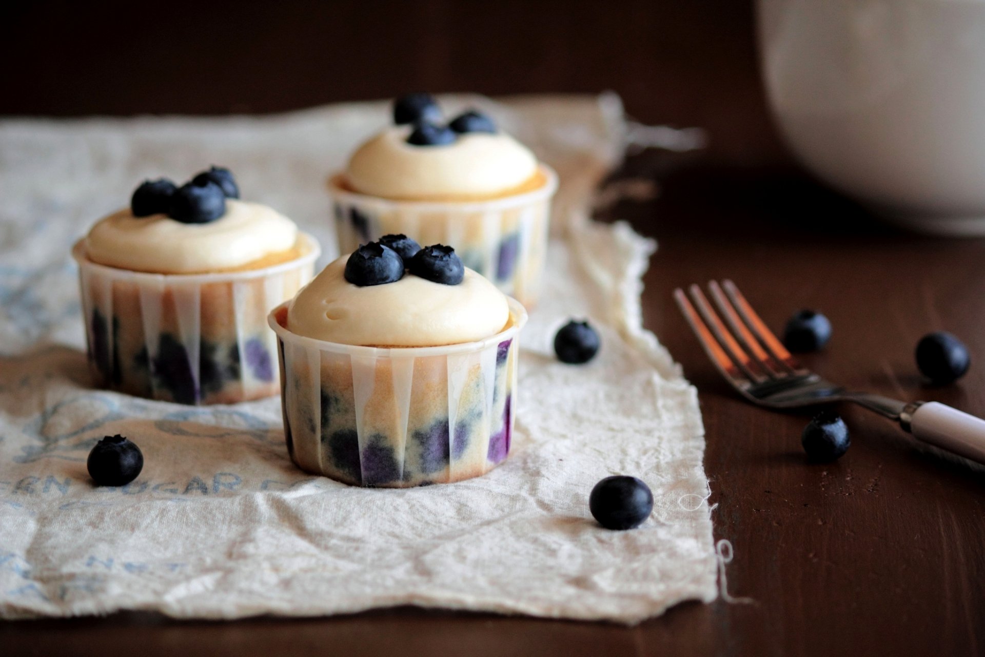Three blueberry topped cupcakes sit on a textured cloth, surrounded by scattered berries and a fork, creating a delightful food-inspired HD wallpaper background.