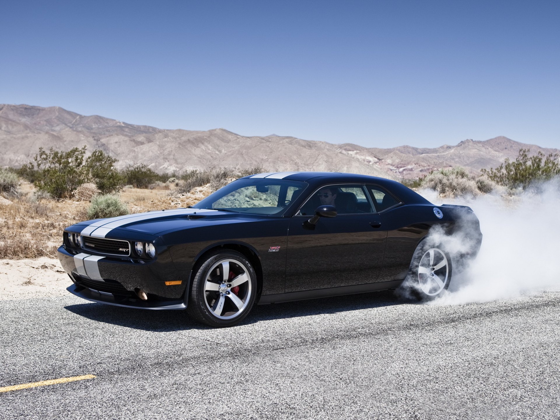 HD desktop wallpaper featuring a black Dodge Challenger performing a burnout on a desert road with mountains in the background.