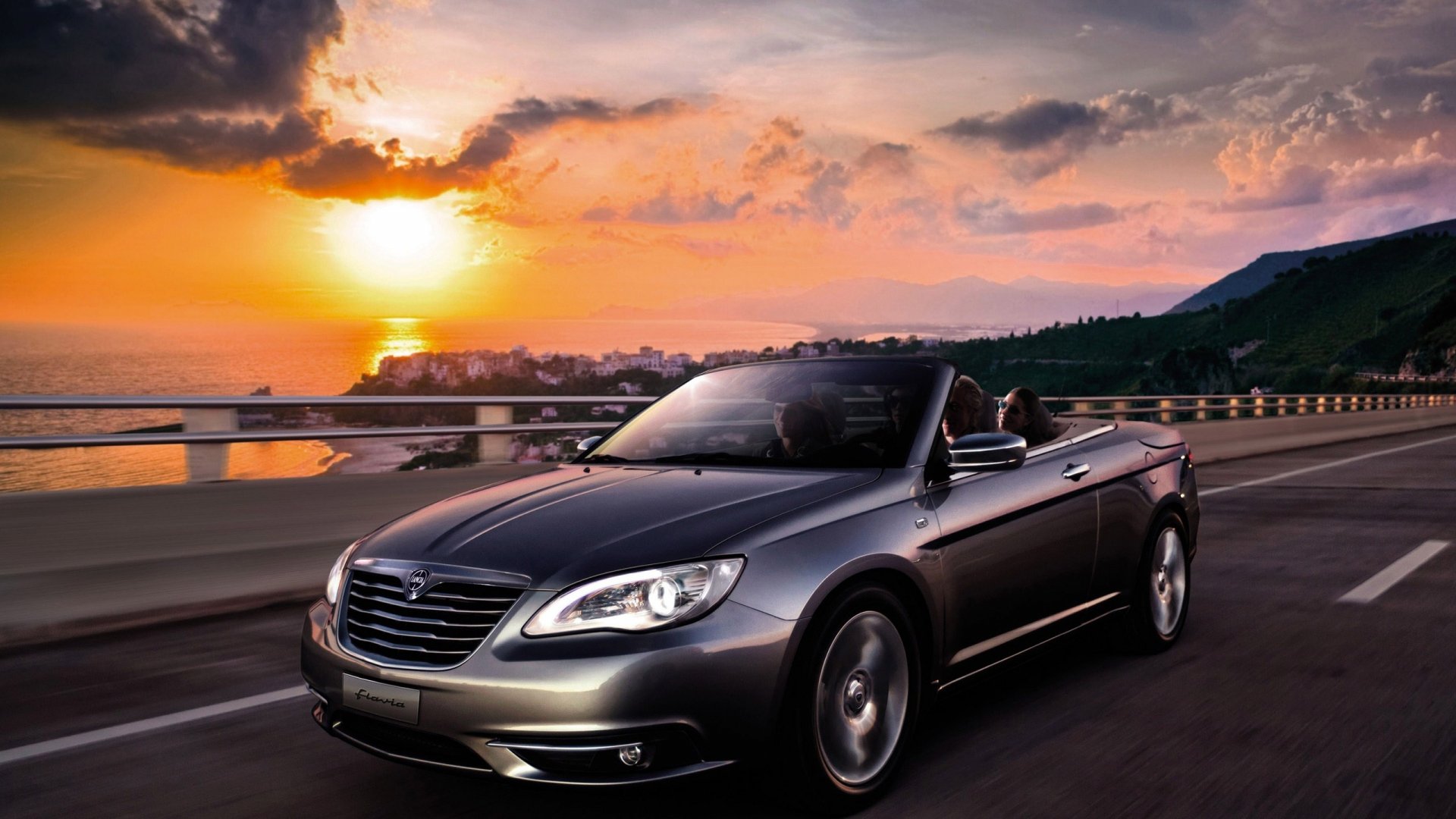 HD desktop wallpaper featuring a sleek Lancia Flavia convertible driving along a scenic coastal road at sunset.