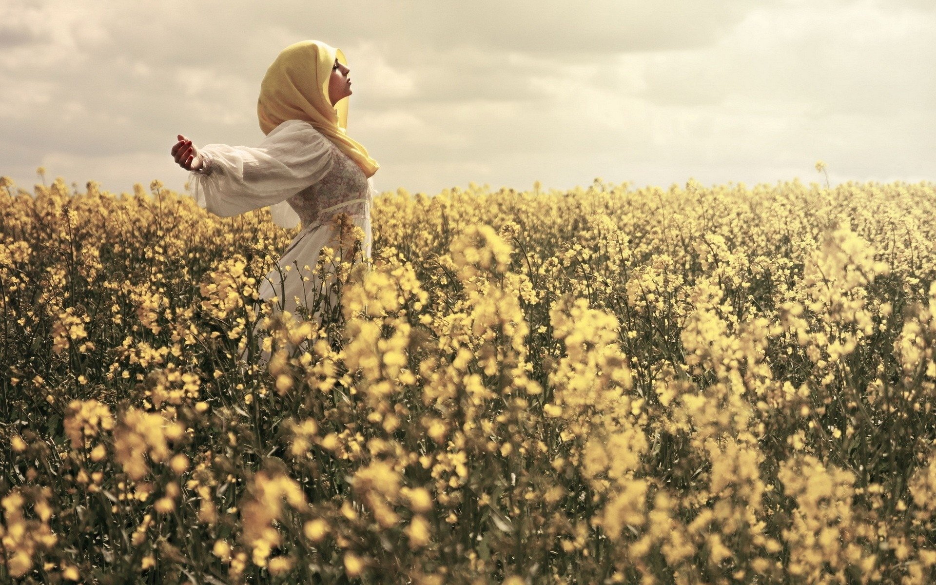 A woman in a yellow hijab stands joyfully in a vibrant field of flowers under a soft sky, capturing a serene mood. This high-definition image serves as an inspiring desktop wallpaper.