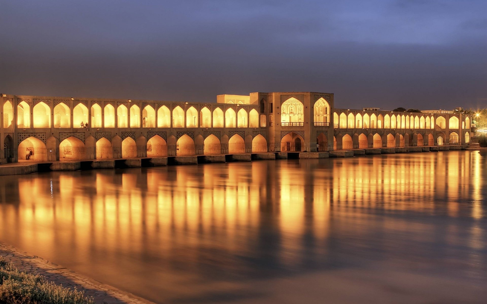 An illuminated Khaju Bridge in Isfahan, Iran, reflects beautifully on the water at dusk, showcasing the elegance of this iconic man-made structure. HD wallpaper background.