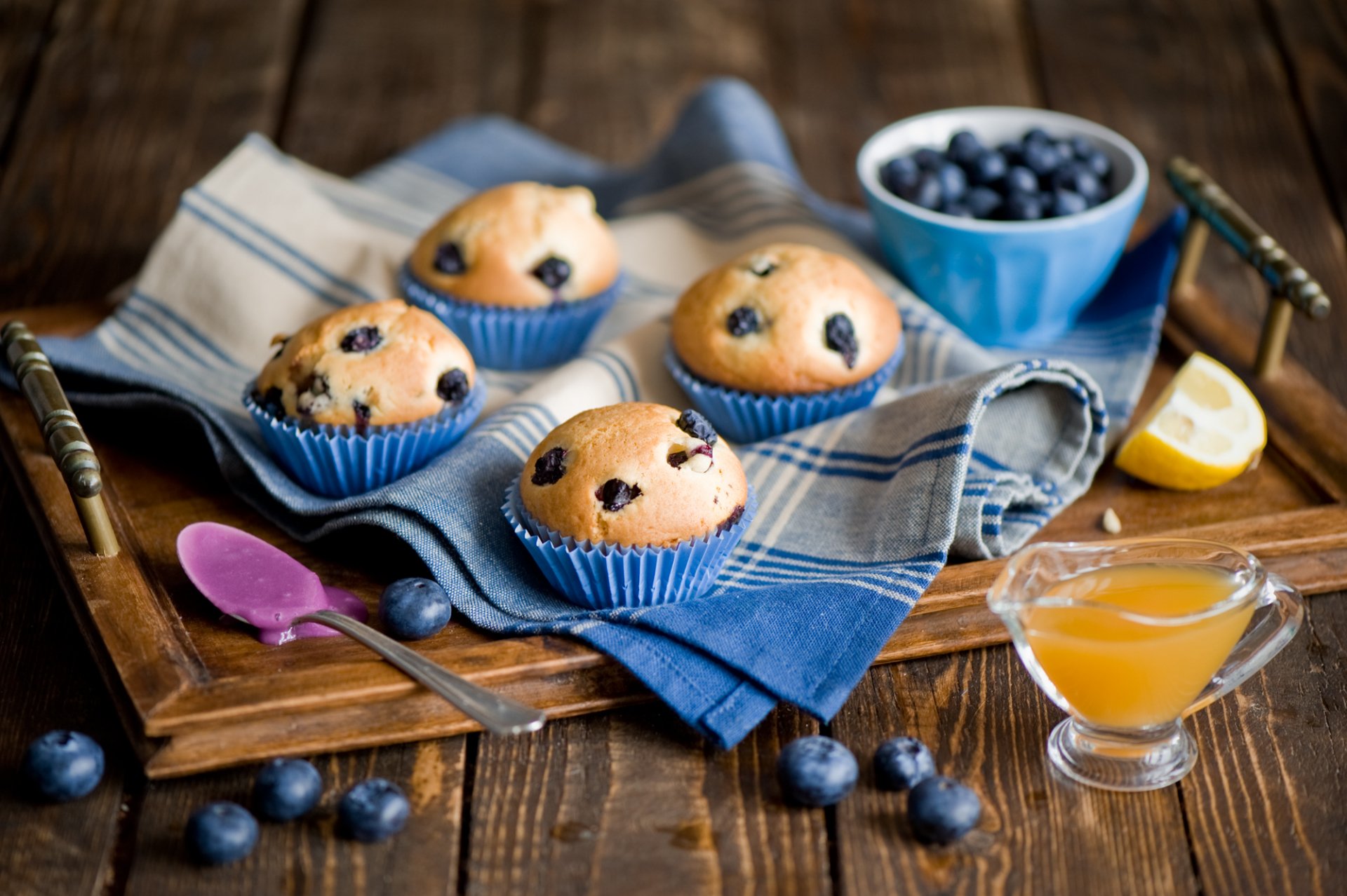 A cozy arrangement of blueberry muffins on a wooden tray with a small bowl of blueberries, a spoon, and a glass of syrup, complementing the inviting atmosphere of sweets and beverages.