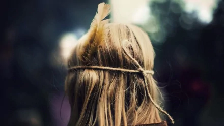 HD desktop wallpaper featuring the back view of a woman with long blonde hair adorned with feathers and a rustic headband against a blurred natural background.