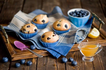 A cozy arrangement of blueberry muffins on a wooden tray with a small bowl of blueberries, a spoon, and a glass of syrup, complementing the inviting atmosphere of sweets and beverages.