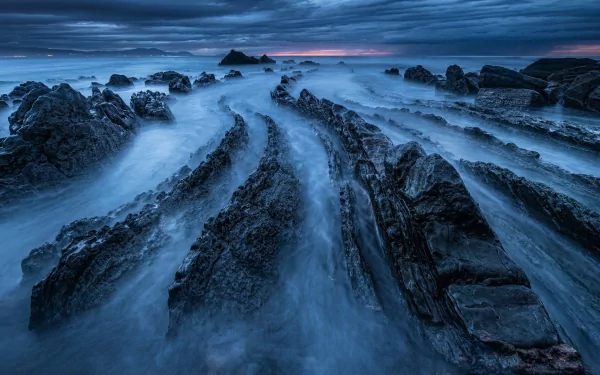 HD PC desktop wallpaper featuring dramatic ocean waves flowing through jagged, rocky formations under a moody, cloud-filled sky at dusk.