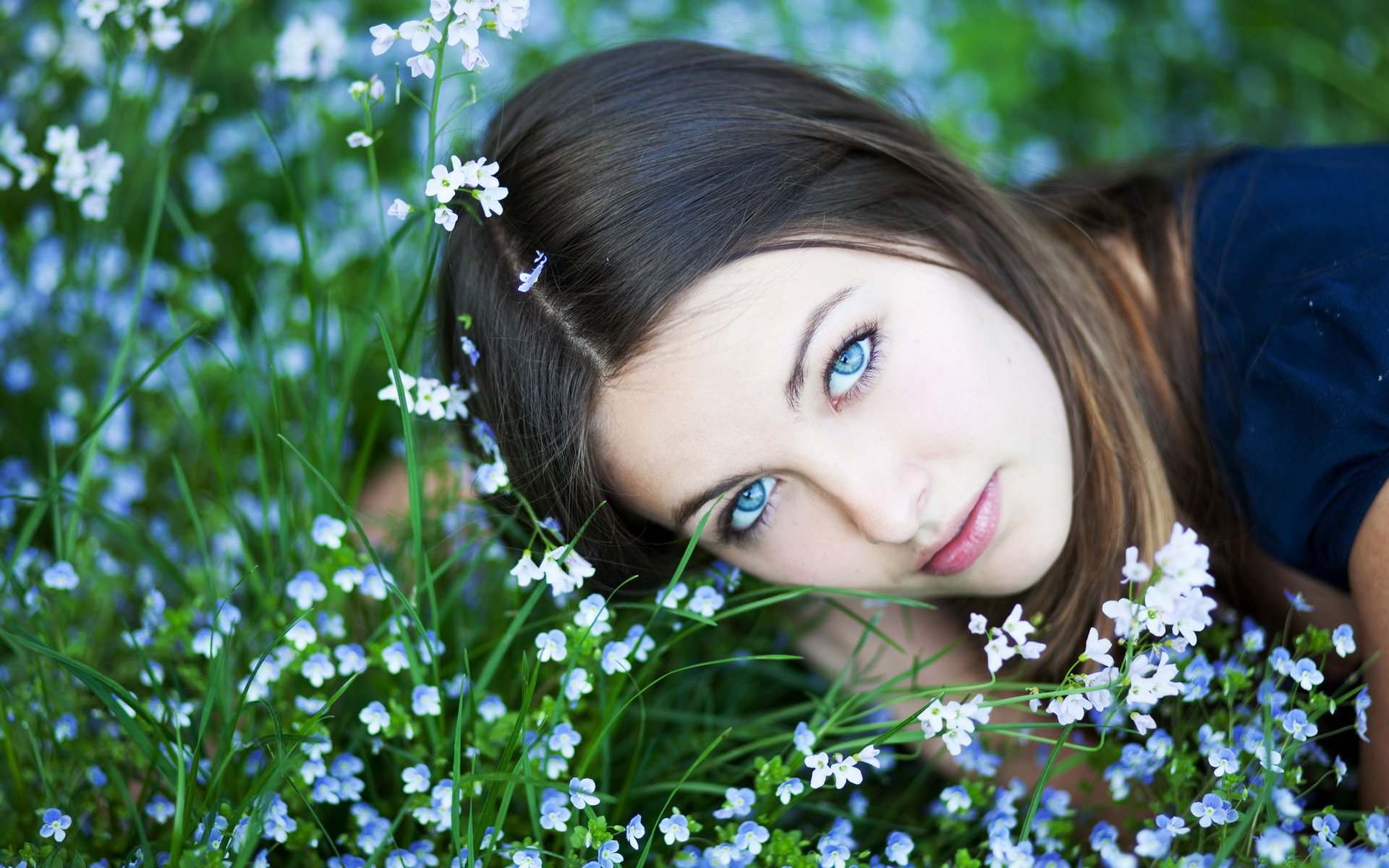 A woman with captivating blue eyes lies amidst a lush field of small white flowers, evoking a serene and contemplative mood. HD desktop wallpaper and background.