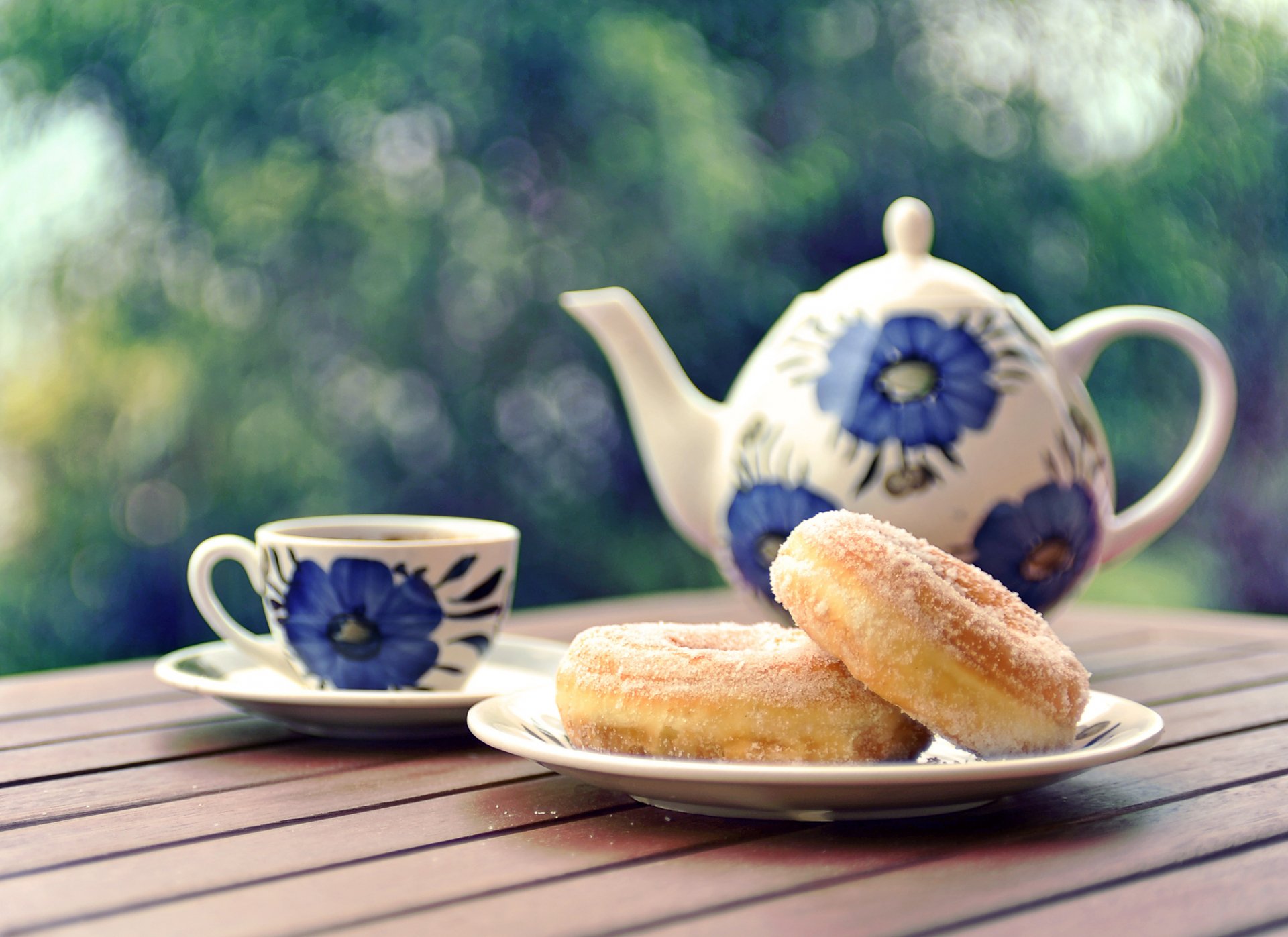 A charming HD desktop wallpaper featuring a decorative teapot and cup, accompanied by two sugary donuts on a plate, set against a serene outdoor backdrop.