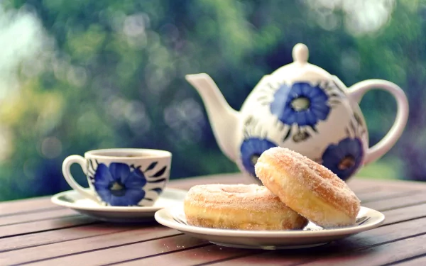 A charming HD desktop wallpaper featuring a decorative teapot and cup, accompanied by two sugary donuts on a plate, set against a serene outdoor backdrop.