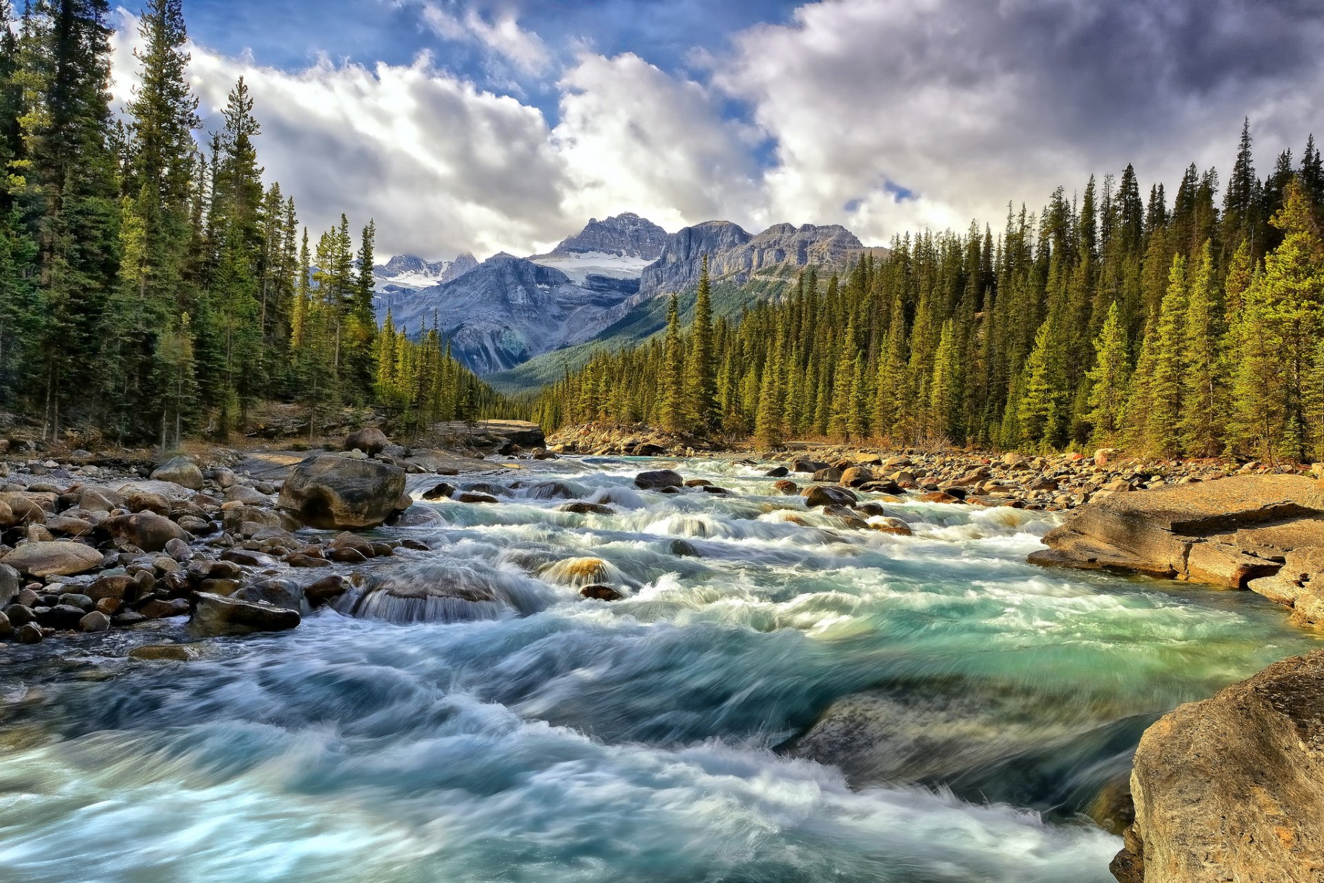 HD PC desktop wallpaper showcasing a vibrant river flowing through a dense forest with mountains under a dramatic cloudy sky.